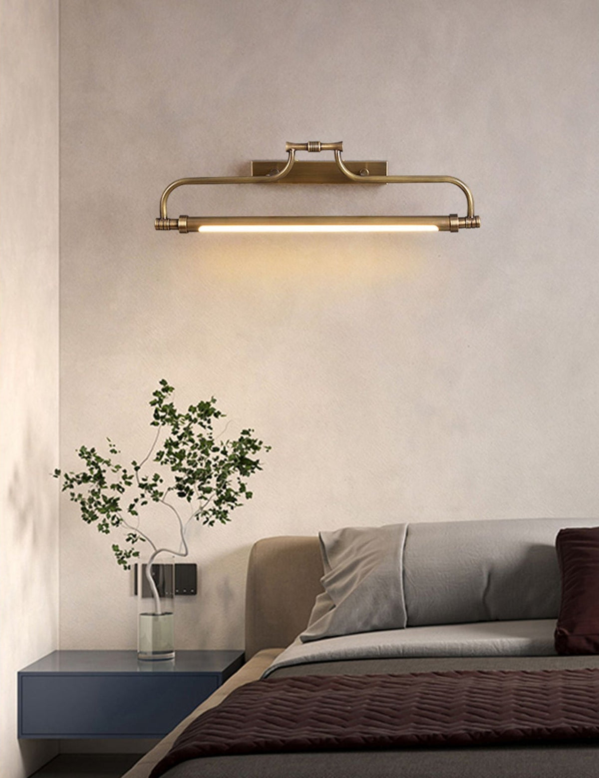Brass wall light from Clérachic above a bed with grey and plum bedding, a navy bedside table and a green sprig in a vase.