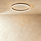 Clérachic slim black round ceiling light with a glowing acrylic ring and pale centre disc, flush on limewash plaster.