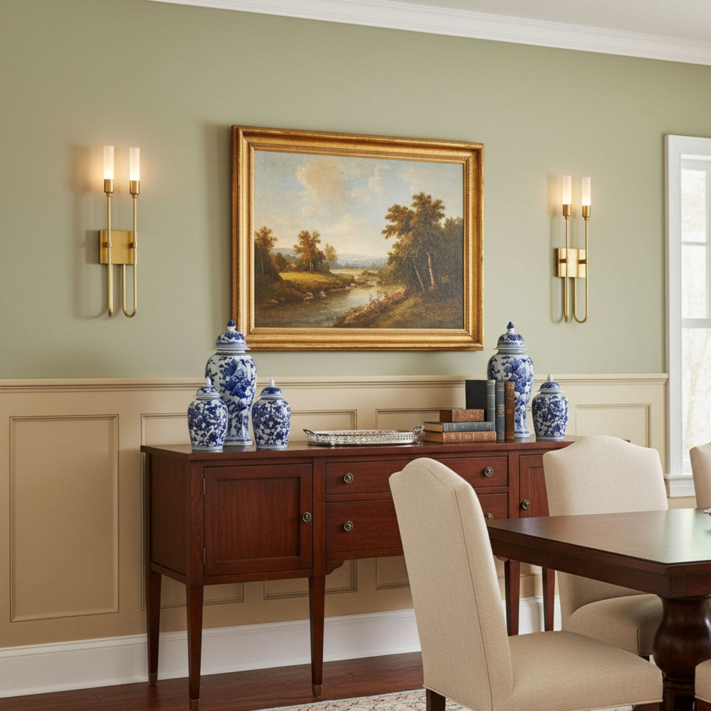 A pair of Clérachic brass wall sconces flanking a gilt-framed landscape painting above a dark wood sideboard with blue-and-white ginger jars.