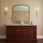 Lit brass Clérachic sconces framing an arched gilt mirror above a mahogany chest of drawers with a silver candelabra, on a cream panelled wall.