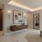 Brass Clérachic wall lights on a pale stone-effect wall by a framed abstract print and a floating walnut unit, under a glowing cove ceiling.