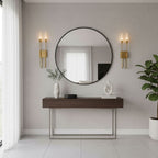 A pair of brass Clérachic two-light sconces beside a large round black-framed mirror above a walnut console, with a leafy plant on the right.