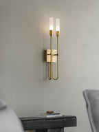 Brass two-light Clérachic sconce on a pale grey wall, lit, with the edge of a dark wooden bench and a small book stack below.
