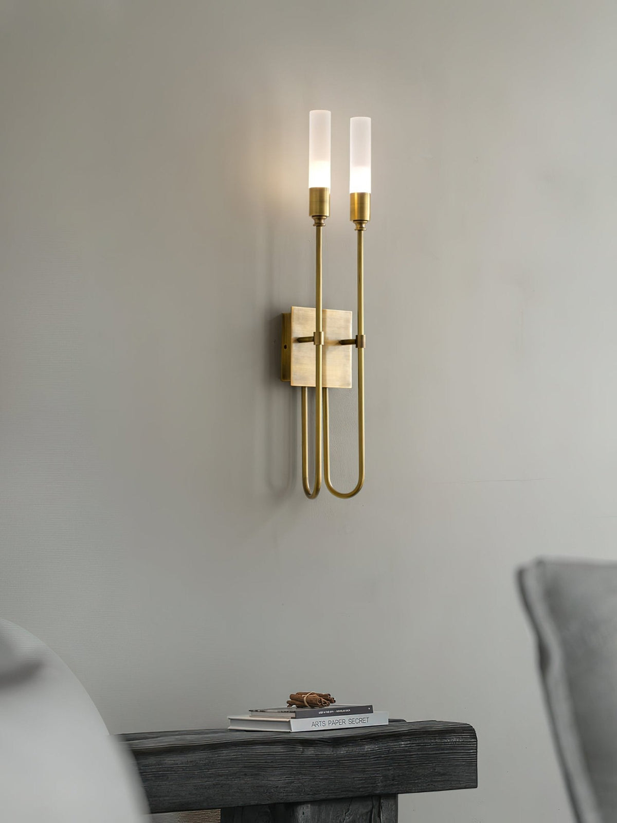 Brass two-light Clérachic sconce on a pale grey wall, lit, with the edge of a dark wooden bench and a small book stack below.