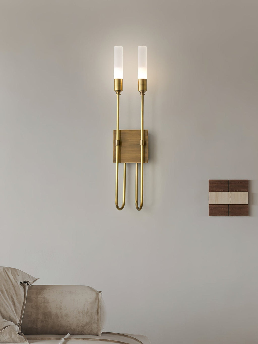 This Clérachic two-light sconce in brass on a soft grey wall, with a sofa corner below and a small wood-and-white panel to the right.