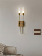 This Clérachic two-light sconce in brass on a soft grey wall, with a sofa corner below and a small wood-and-white panel to the right.