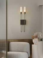 Black Clérachic two-light sconce lit on a grey wall above a boucle armchair and a white plinth holding dried stems and a sculpted vase.