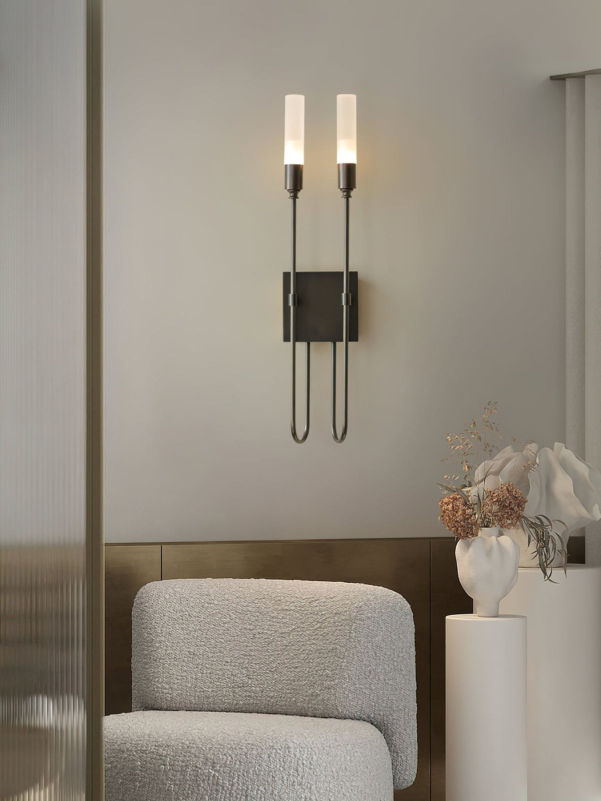 Black Clérachic two-light sconce lit on a grey wall above a boucle armchair and a white plinth holding dried stems and a sculpted vase.