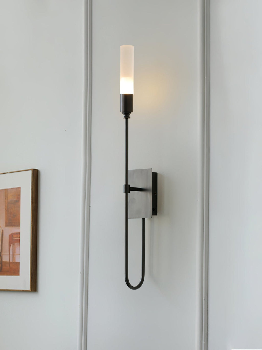 Single-light black Clérachic sconce lit on a white panelled wall, with the edge of a framed picture beside the tall frosted tube.