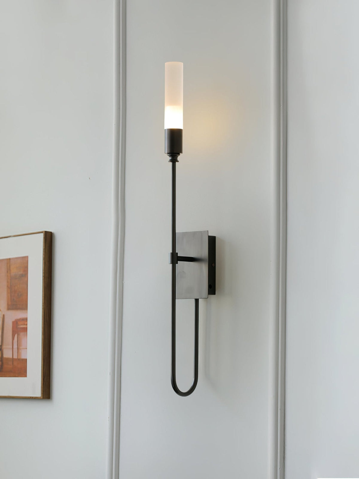 Single-light black Clérachic sconce lit on a white panelled wall, with the edge of a framed picture beside the tall frosted tube.