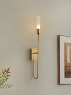 Single-light brass Clérachic sconce glowing on a warm wall, with a framed picture on the right and green leaves at the lower left.