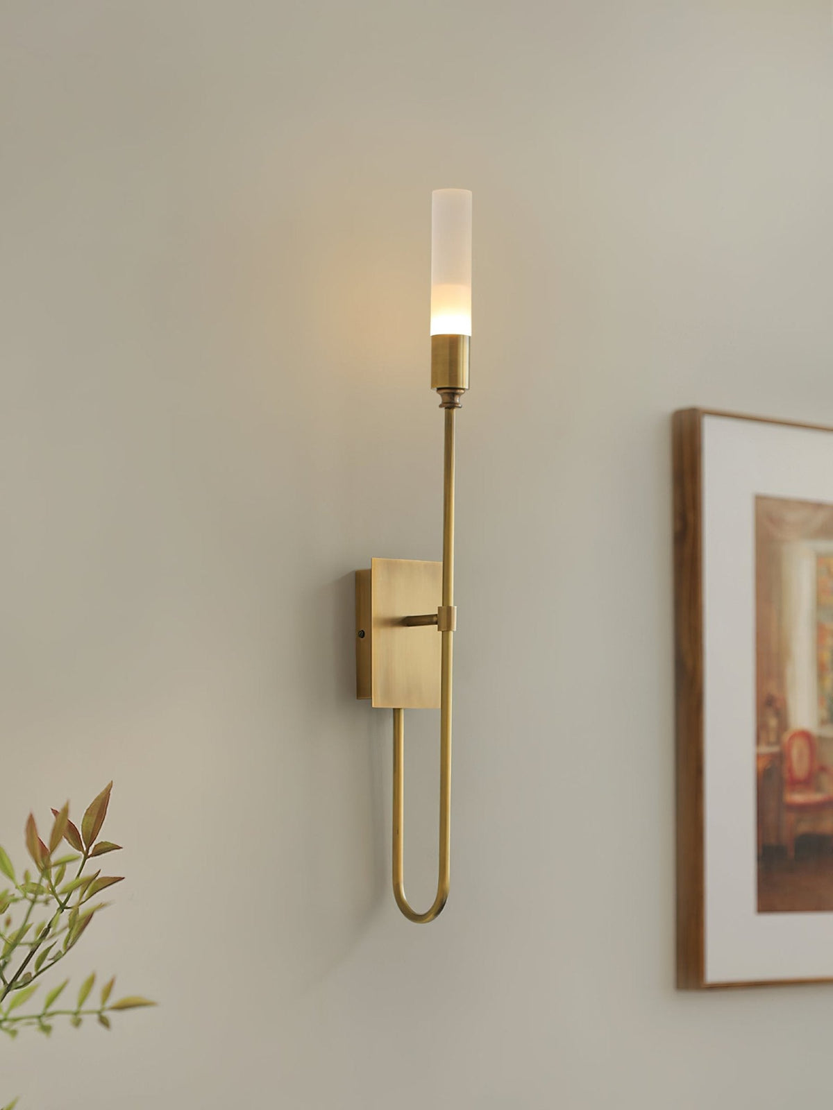 Single-light brass Clérachic sconce glowing on a warm wall, with a framed picture on the right and green leaves at the lower left.