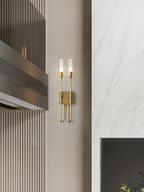 Small brass two-light Clérachic sconce on a pale panelled wall beside a marble-look surface and a dark vent, shown from a distance.