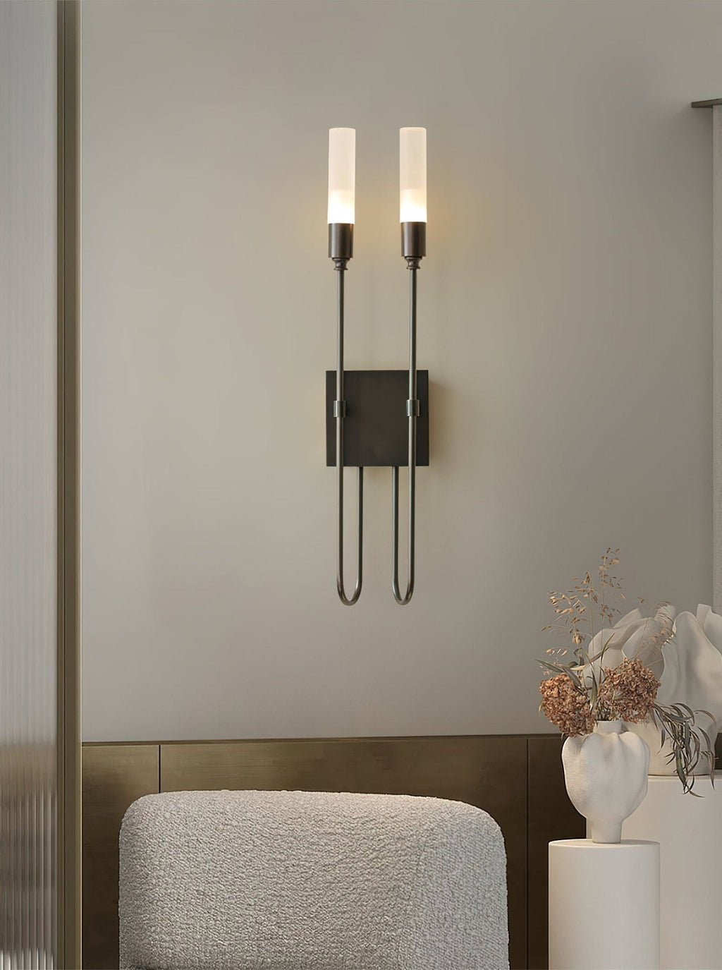 Black two-light Clérachic sconce on a grey wall above a boucle armchair, with a white vase of dried flowers at the right.
