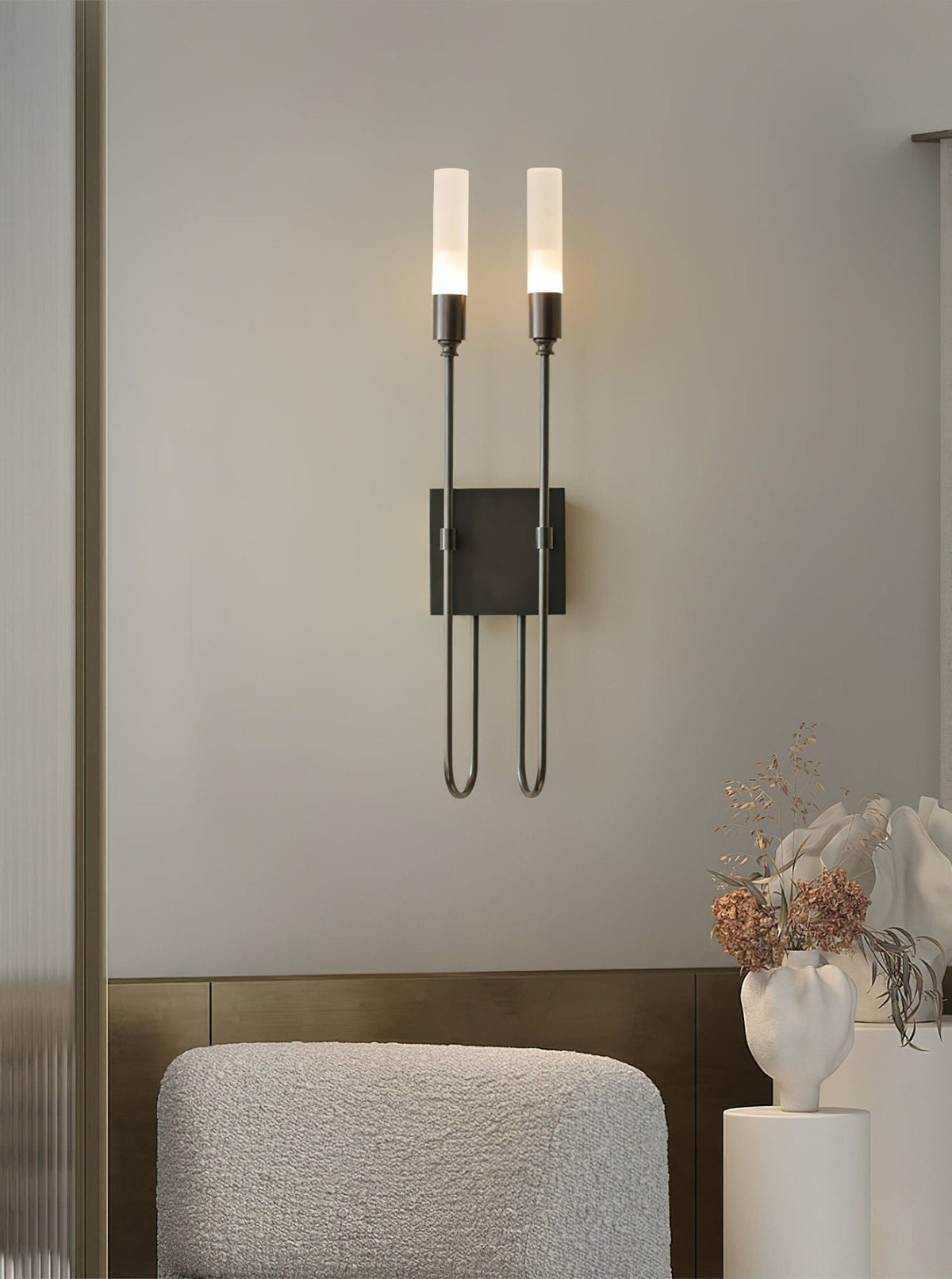 Black two-light Clérachic sconce on a grey wall above a boucle armchair, with a white vase of dried flowers at the right.
