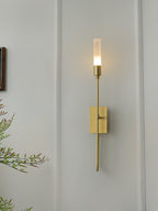 Single-light brass Clérachic sconce on a pale panelled wall, its frosted tube glowing, with bamboo-like leaves at the left edge.