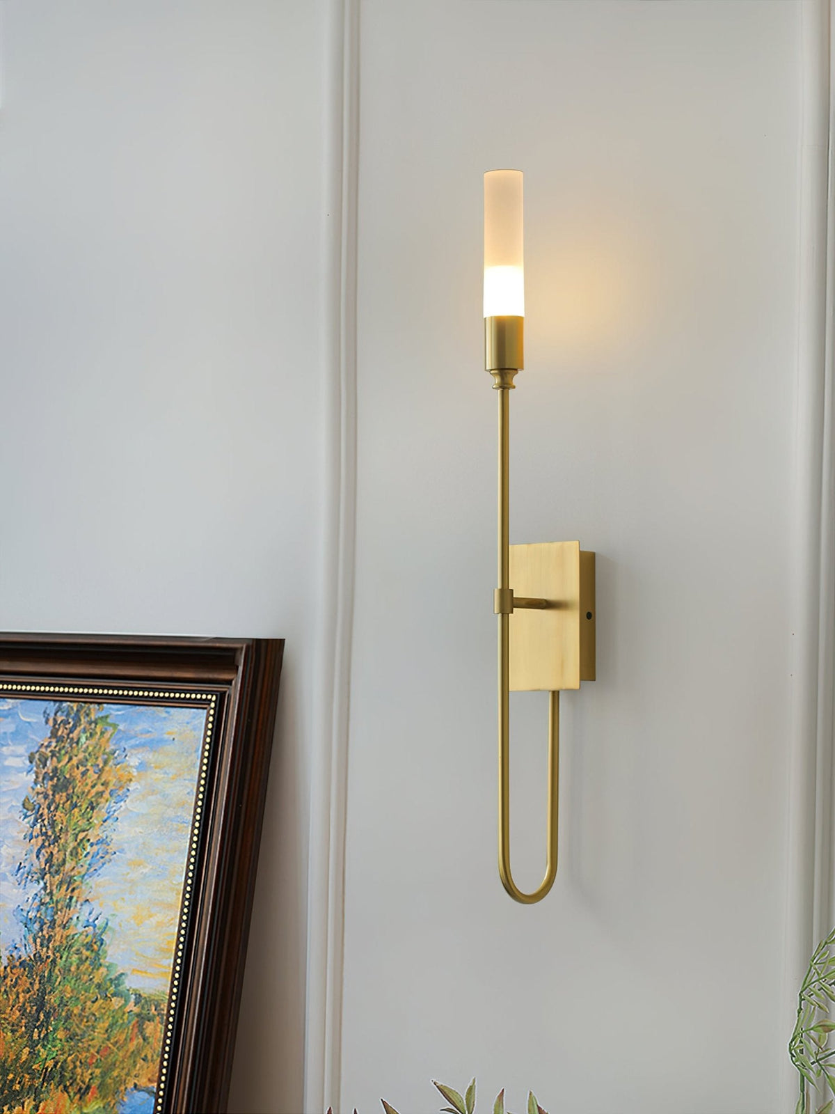 Single-light brass Clérachic sconce with a looped lower arm on a panelled wall, beside a framed autumn-tree painting.