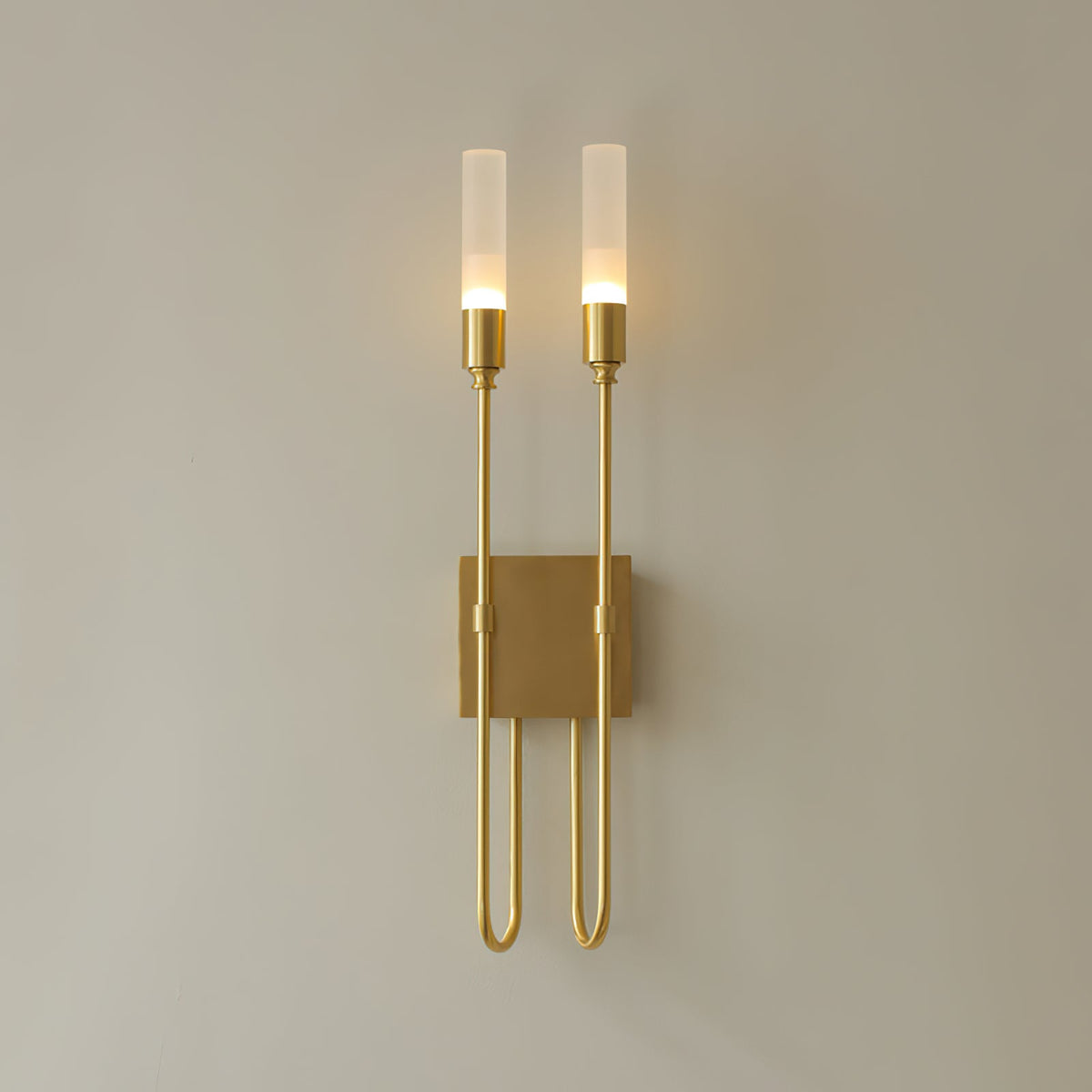 Straight-on view of this Clérachic two-light sconce in brass, lit, on a plain warm-grey wall with long U-shaped arms below the plate.