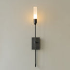 Single-light black Clérachic sconce on a grey wall, its frosted tube glowing warm above a rectangular plate and a curved lower arm.