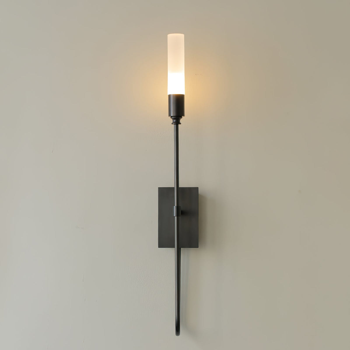 Single-light black Clérachic sconce on a grey wall, its frosted tube glowing warm above a rectangular plate and a curved lower arm.