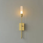 Single-light brass Clérachic sconce glowing on a grey wall, a halo of warm light around the frosted tube.