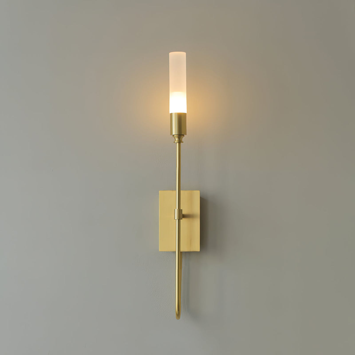 Single-light brass Clérachic sconce glowing on a grey wall, a halo of warm light around the frosted tube.