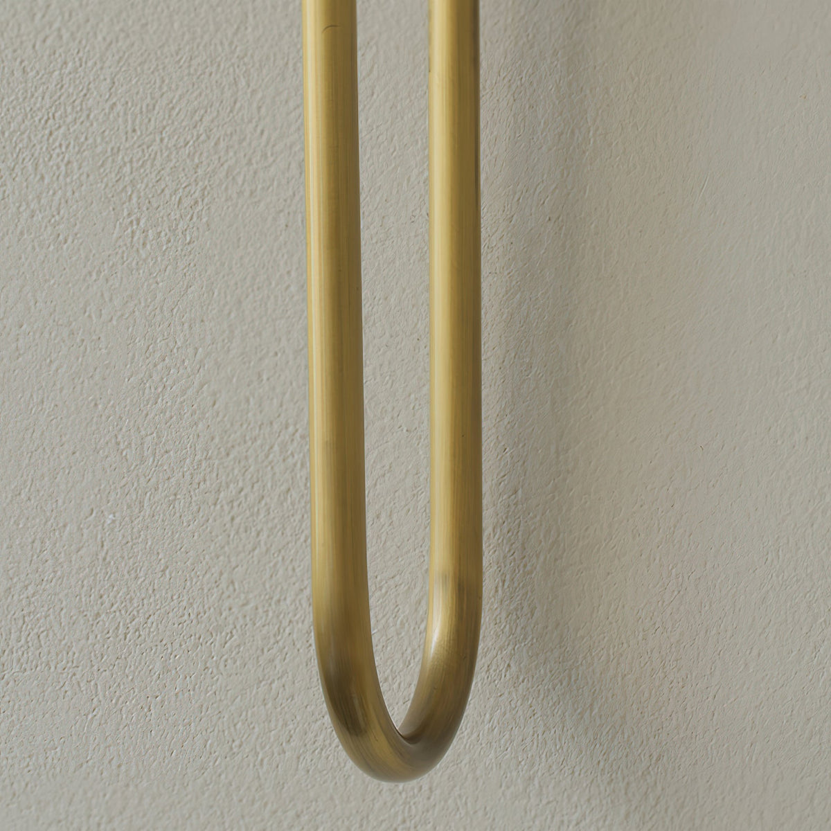 Close view of the rounded U-bend at the base of a brass arm on this Clérachic sconce, against a textured grey wall.
