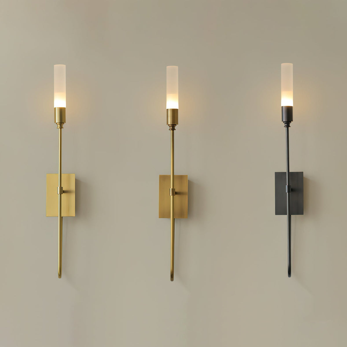 Three single-light Clérachic sconces side by side on a taupe wall, in brass, antique brass and black, each lit.