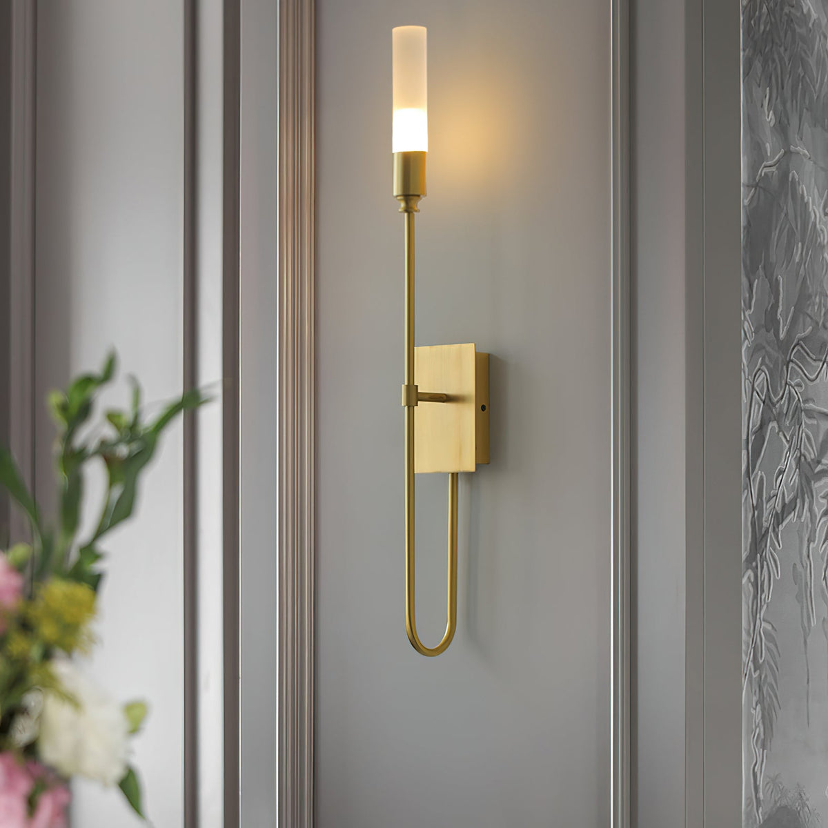 Single-light brass Clérachic sconce on a grey panelled wall, with pink and yellow flowers blurred in the foreground at the left.