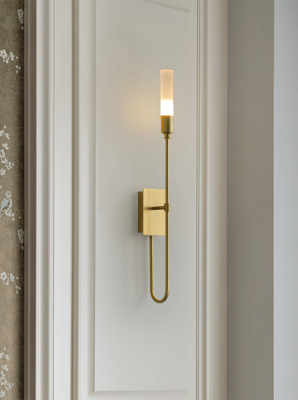 Single-light brass Clérachic sconce on a white panelled wall with a looped lower arm, beside a patterned curtain edge.