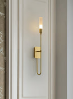 Single-light brass Clérachic sconce on a white panelled wall with a looped lower arm, beside a patterned curtain edge.