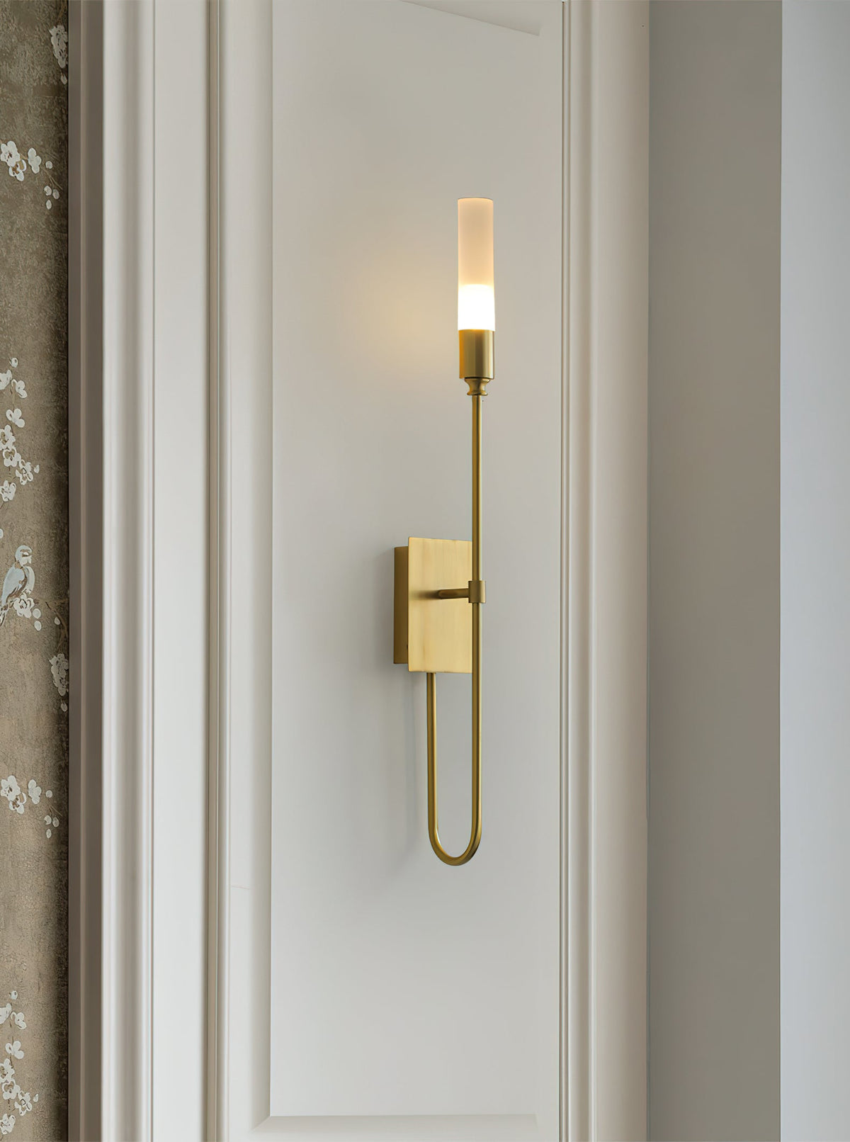 Single-light brass Clérachic sconce on a white panelled wall with a looped lower arm, beside a patterned curtain edge.