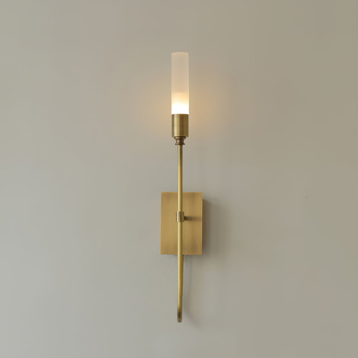 Single-light brass Clérachic sconce lit on a warm grey wall, with a thin arm ending in a small curl below the plate.
