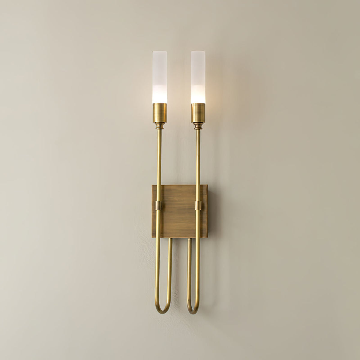 Two-light Clérachic sconce in antique brass on a warm neutral wall, both frosted tubes lit above a small square plate.
