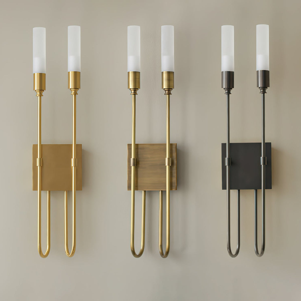 Three Clérachic two-light sconces in brass, antique brass and black, hung side by side on a beige wall with the tubes lit.