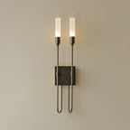 Black two-light Clérachic sconce on a beige wall, with both frosted tubes lit and thin arms dropping to a looped base.