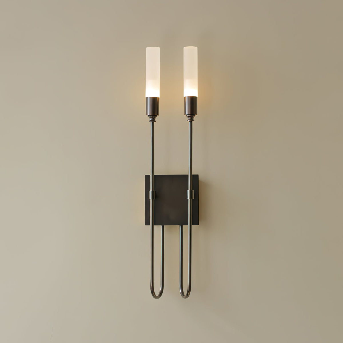 Black two-light Clérachic sconce on a beige wall, with both frosted tubes lit and thin arms dropping to a looped base.