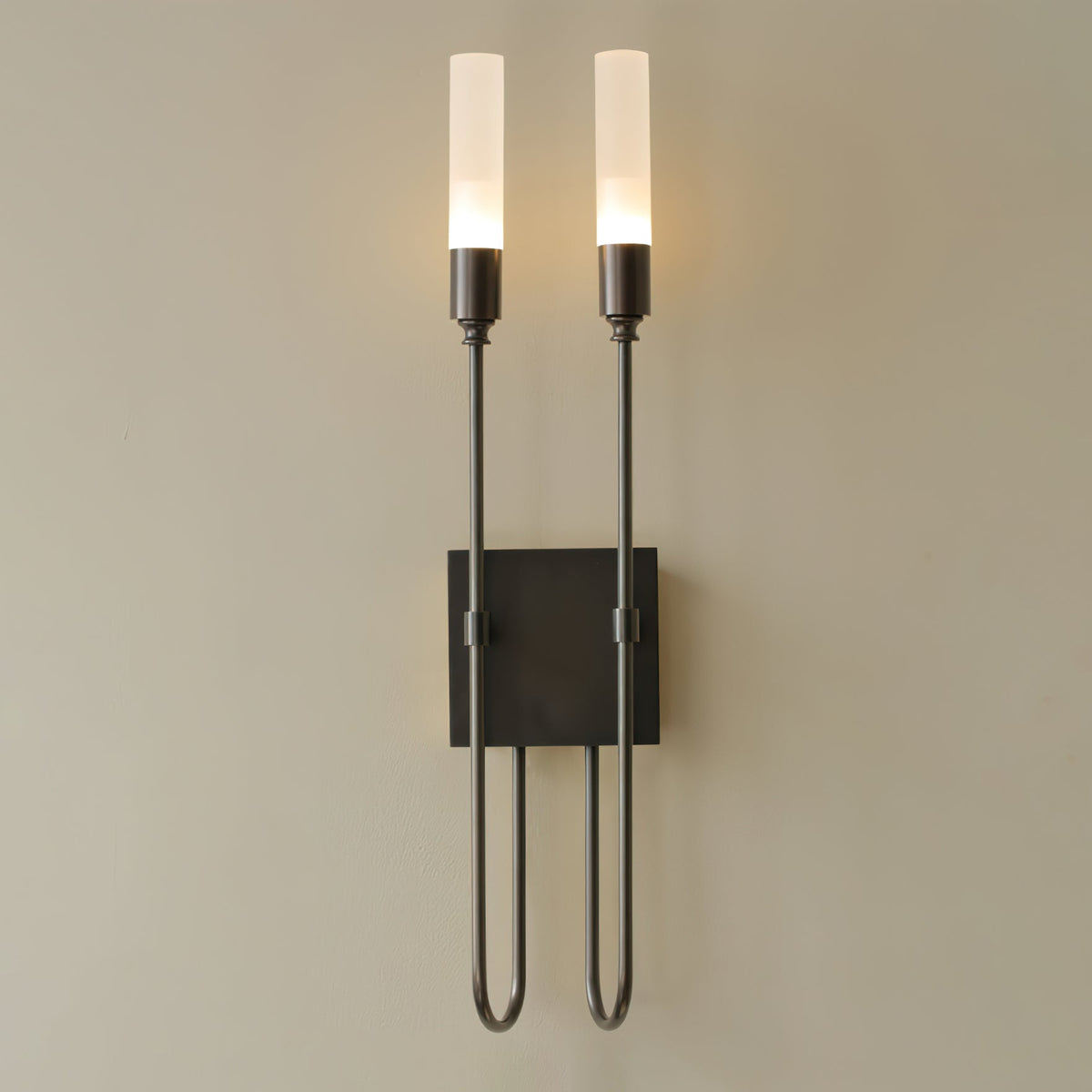 Tall view of this black Clérachic two-light sconce, lit, with its square plate and U-shaped arms on a beige wall.