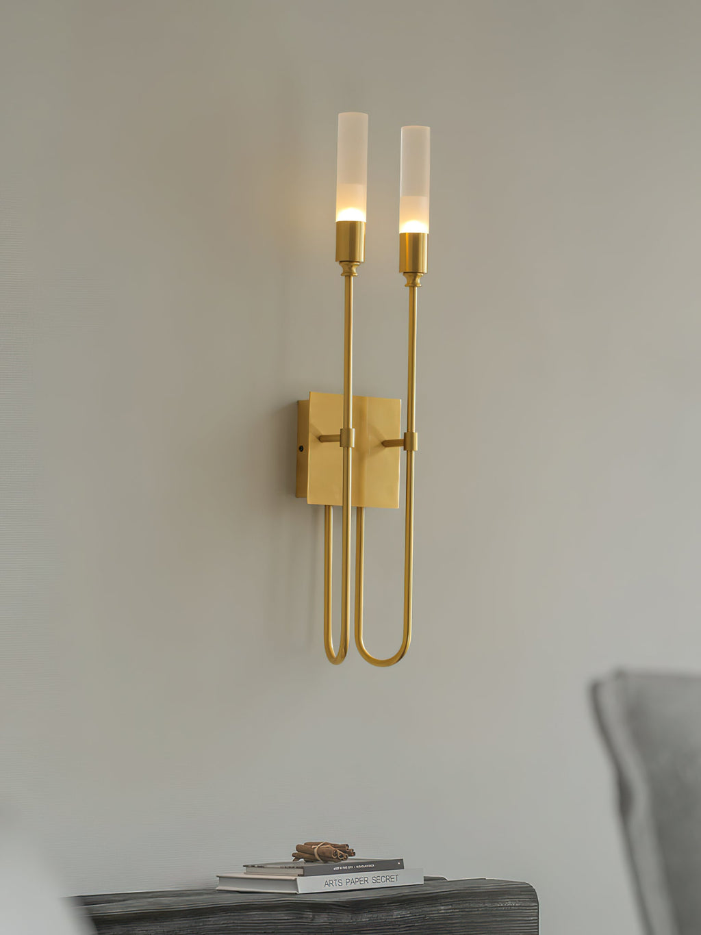 Brass two-light Clérachic sconce on a pale grey wall, with a dark bench, a small book and a grey cushion below.