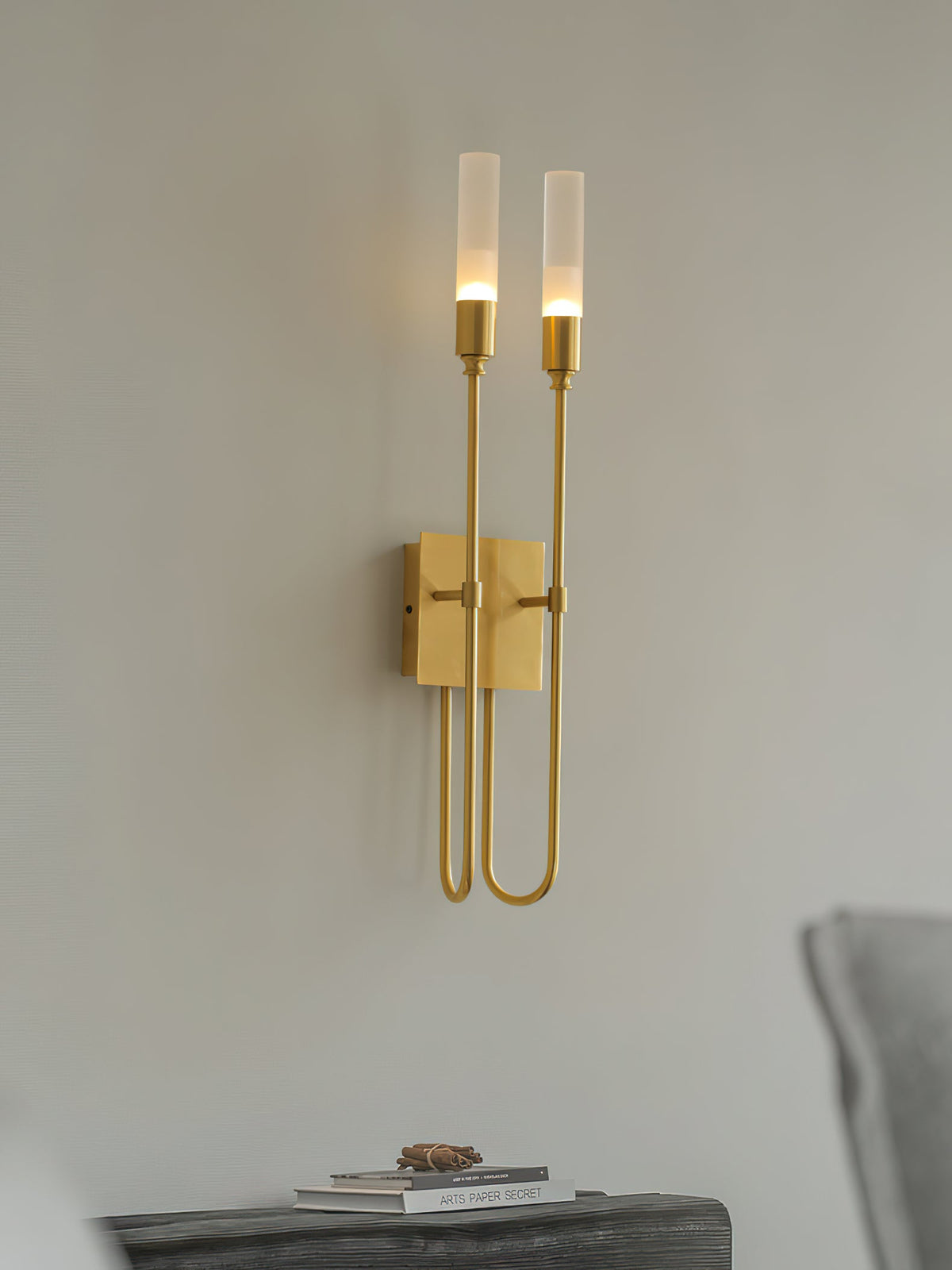 Brass two-light Clérachic sconce on a pale grey wall, with a dark bench, a small book and a grey cushion below.