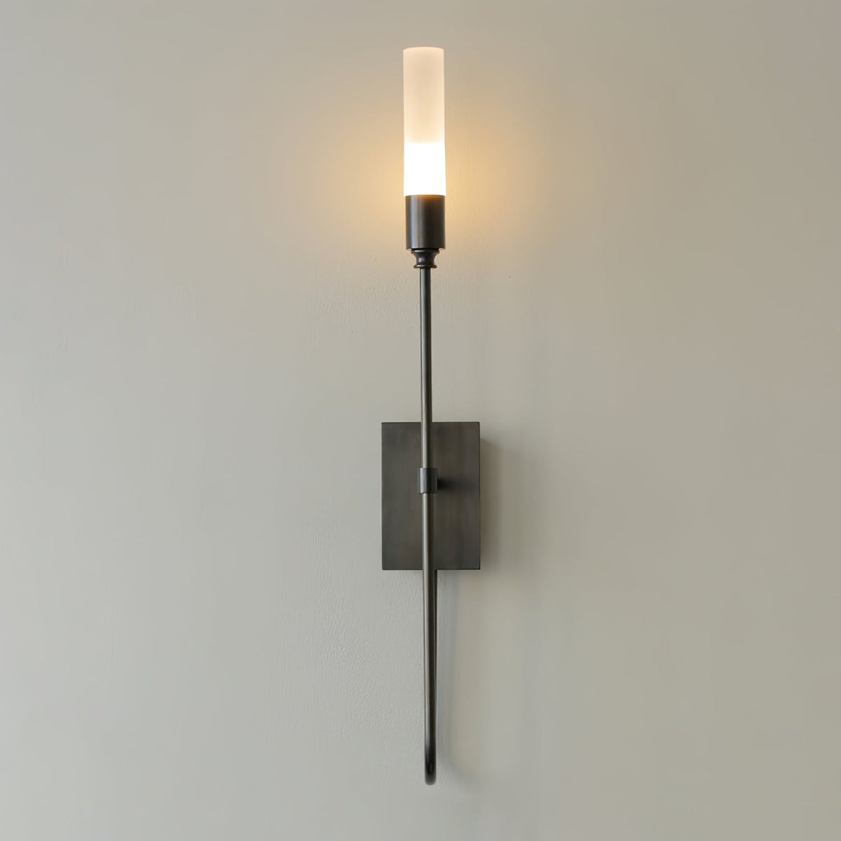 Single-light black Clérachic sconce on a grey wall, its frosted tube glowing above a small dark rectangular plate.