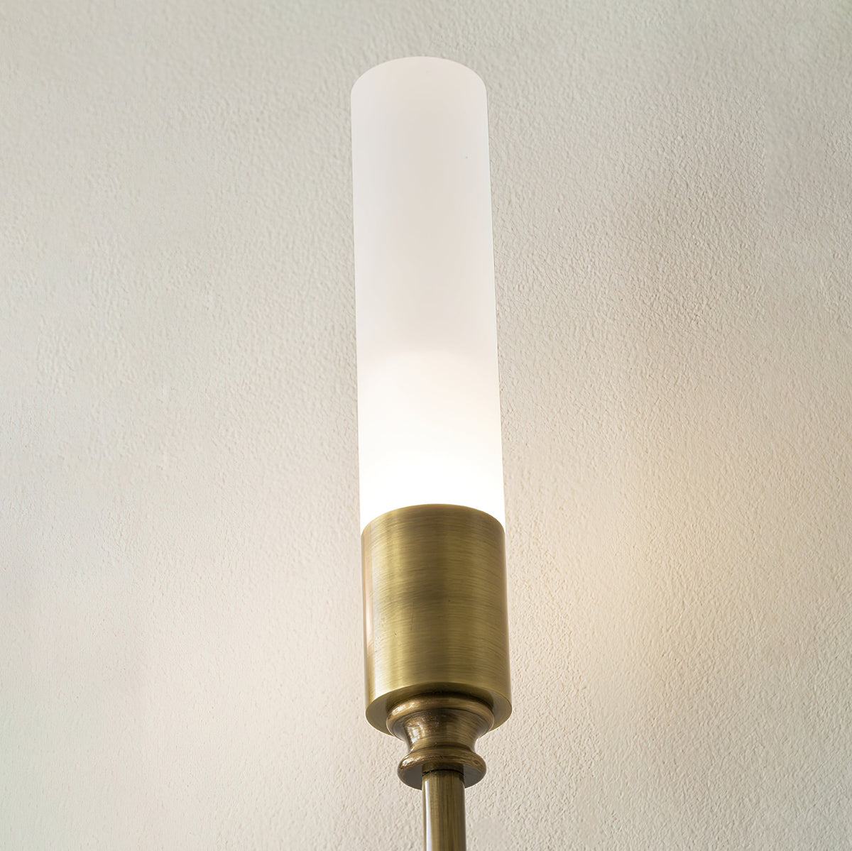 Close view of the frosted tube and aged brass cap on this Clérachic sconce, lit, against a pale wall.