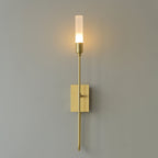 Single-light brass Clérachic sconce with a glowing frosted tube on a grey wall, a long straight arm below the plate.