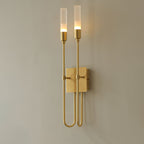 Brass two-light Clérachic sconce, lit, on a grey wall, with arms looping down and back up either side of a square plate.