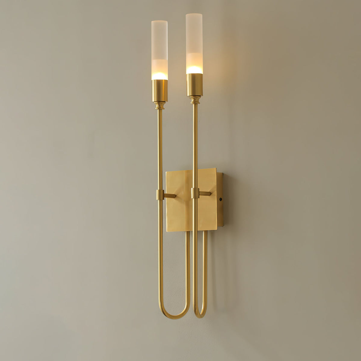 Brass two-light Clérachic sconce, lit, on a grey wall, with arms looping down and back up either side of a square plate.