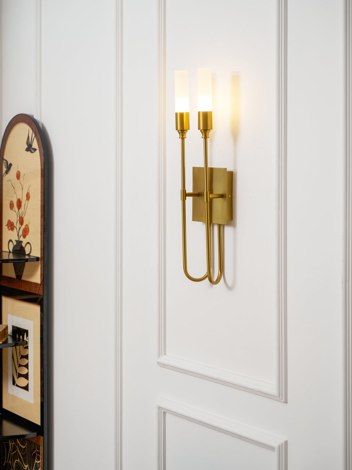 Brass two-light Clérachic sconce on a white panelled wall beside an arched painted cabinet, with the tubes softly lit.