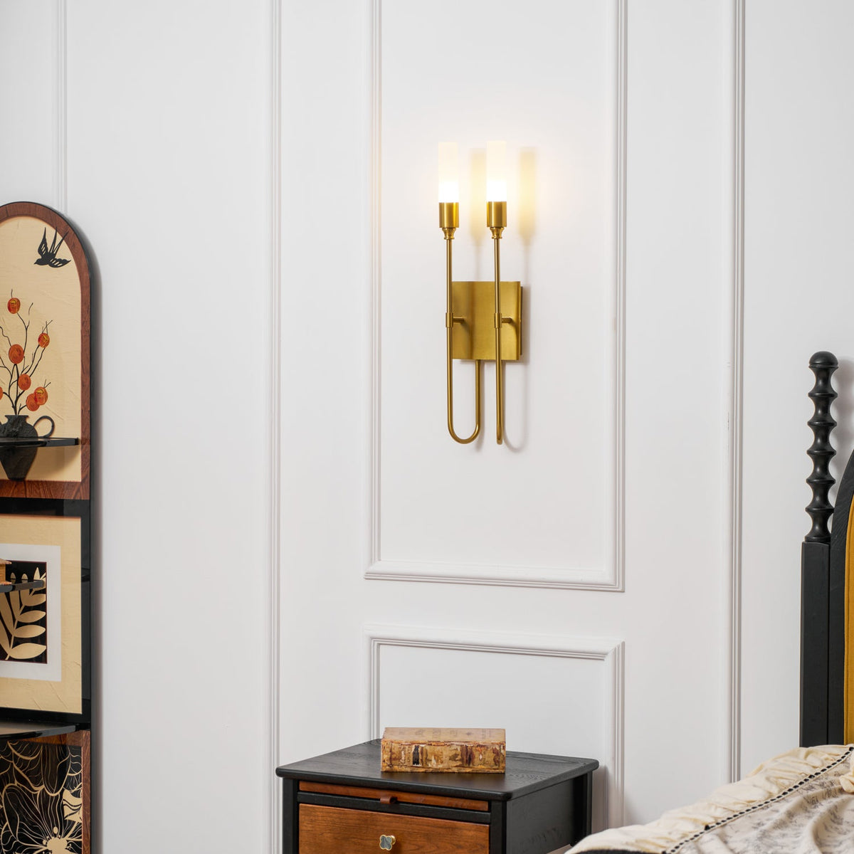 Lit brass two-light Clérachic sconce on a white panelled wall above a black nightstand, with a painted arched cabinet to the left.