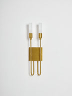 Brass two-light Clérachic sconce, unlit, on a pale grey backdrop, with frosted tubes and looped arms around a small plate.
