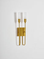 Slightly angled view of this Clérachic two-light sconce in brass on a plain pale wall, with the frosted tubes unlit.