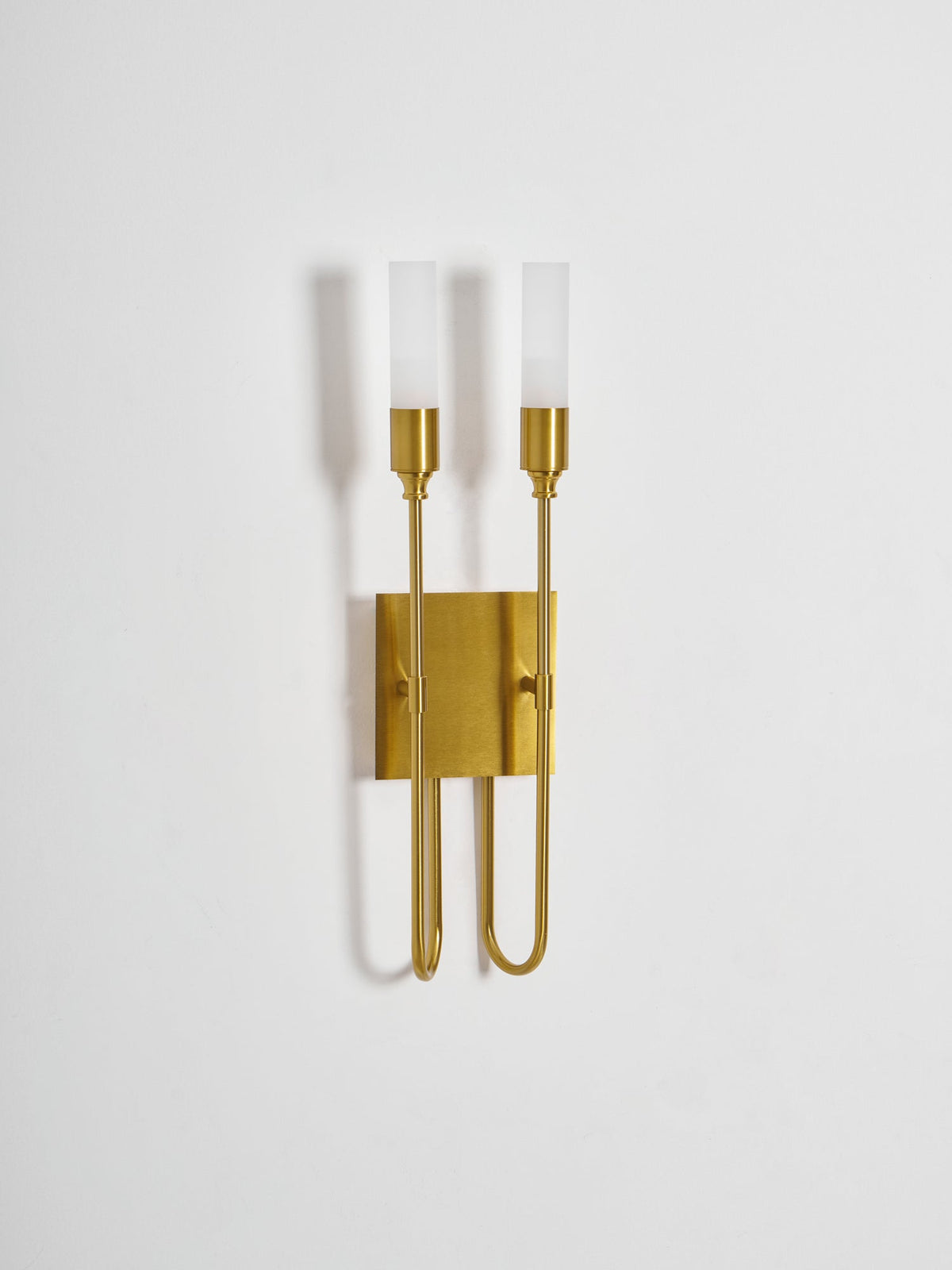 Slightly angled view of this Clérachic two-light sconce in brass on a plain pale wall, with the frosted tubes unlit.